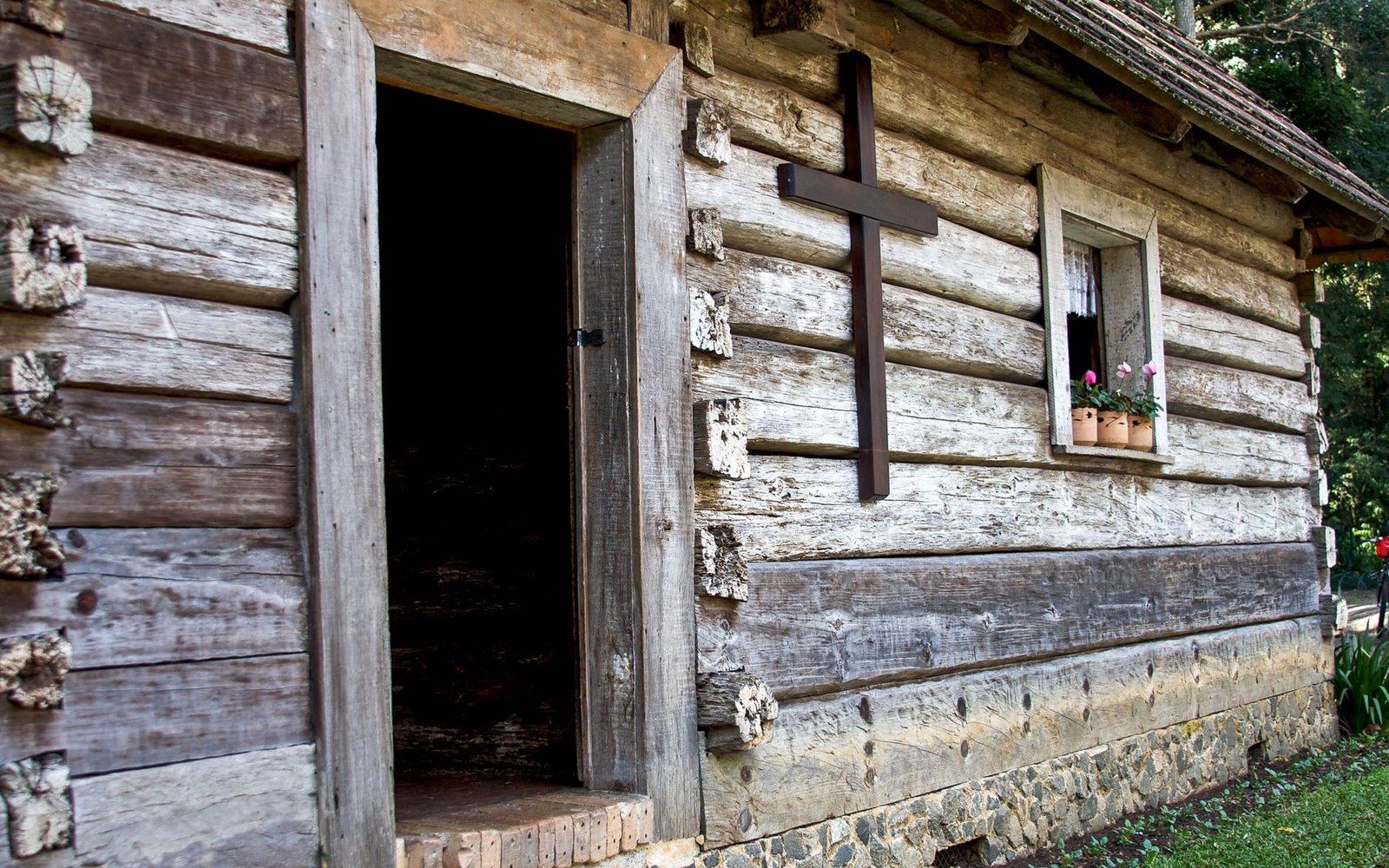 Weathered log cabin chapel with a wooden cross and flower-lined window beside an open door