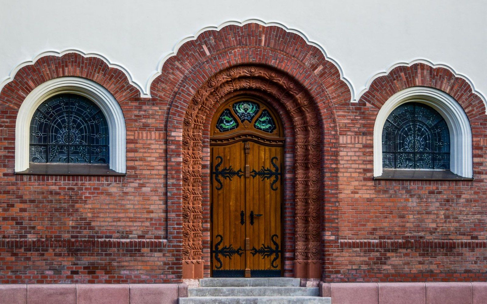 Arched wooden door with wrought-iron hinges flanked by round windows in a brick facade